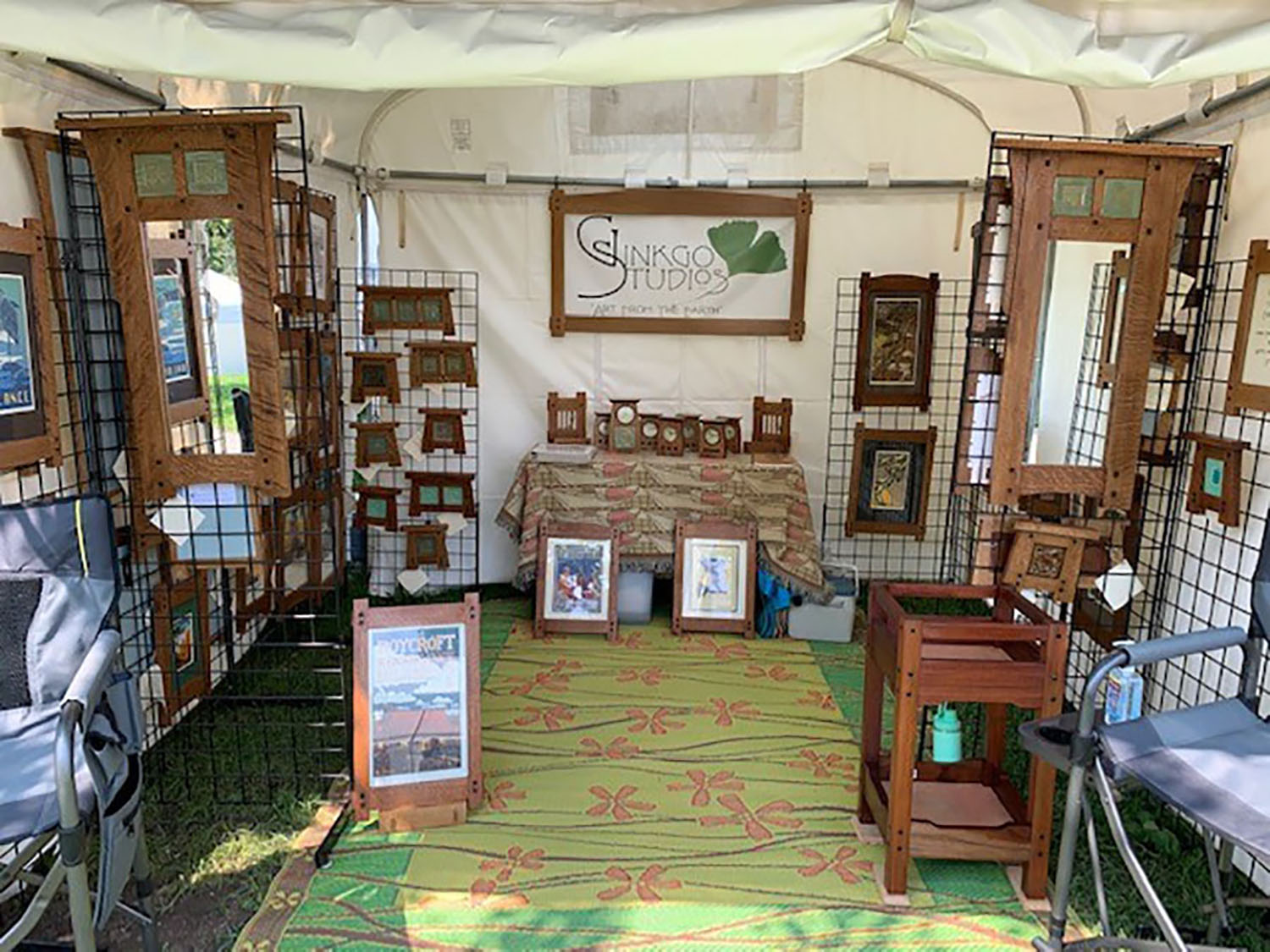 Art fair booth displaying framed mirrors, clocks, and wooden crafts by Slinko Studios, arranged on wire grids and tables.